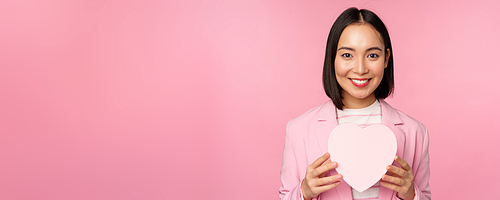 This is for you. Romantic cute asian corporate woman, girl in suit, showing heart-shaped box with gift, standing over pink background.