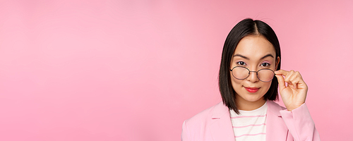 Portrait of asian businesswoman in glasses, looking intrigued at camera and smiling, professional saleswoman staring with interest, pink background.