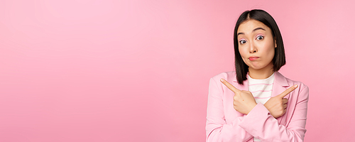 Decision concept. Young asian businesswoman, corporate worker pointing fingers sidways, two directions, showing variants, looking indecisive while choosing, pink background.