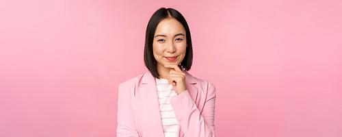 Image of asian businesswoman standing in thinking pose, brainstoming, wearing suit. Korean saleswoman, entrepreneur posing against pink background.