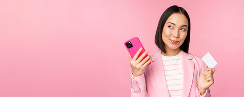 Smiling asian corporate woman, lady in suit thinking, holding smartphone and credit card, plan to buy smth online, shopping with mobile phone, pink background.