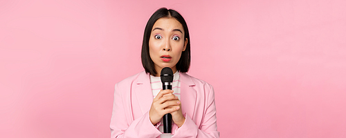 Young asian saleswoman, office lady with suit, holding microphone and looking shocked at camera, talking, giving speech, standing over pink background. Copy space