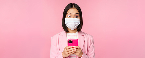 Business people and covid-19 concept. Young asian businesswoman in suit and medical face mask, using smartphone app, standing over pink background.