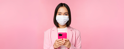 Business people and covid-19 concept. Japanese corporate office lady in suit and medical face mask, using mobile phone and smiling at camera, pink background.