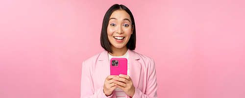 Portrait of asian girl in suit with smartphone, smiling and looking happy, standing over pink studio background. Copy space