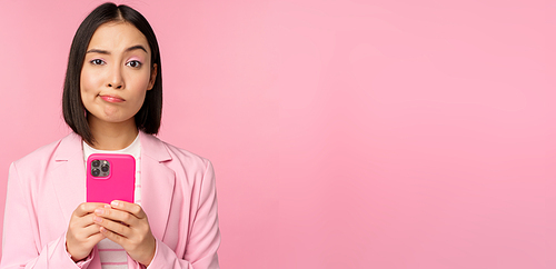 Doubtful businesswoman in suit, holding smartphone, grimacing, making skeptical face expression, standing over pink background.