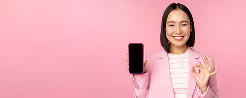 Satisfied smiling asian businesswoman recommending mobile phone app, website company on smartphone, showing screen and okay sign, pink background.