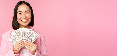 Finance, microcredit and people concept. Happy smiling asian businesswoman showing dollars money, standing in suit against pink background.