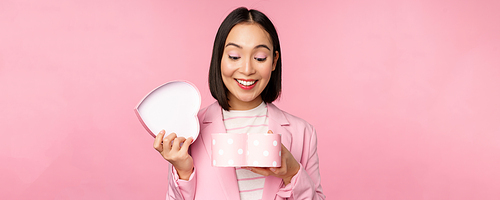 Happy cute korean girl in suit, opens up heart shaped box with romantic gift on white day holiday, standing in suit over pink background.