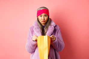 Upset senior asian woman open shopping bag and looking sad at camera, standing over pink background.