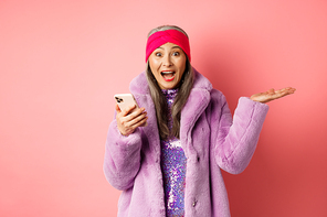 Online shopping and fashion concept. Woman checking out awesome promo offer on smartphone, scream of joy at camera, looking with surprised and happy face, pink background.
