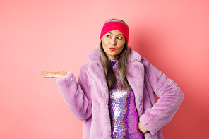 Fashion and shopping concept. Stylish asian senior woman in purple winter outfit demonstrating product on hand. Cool old lady in trendy clothes holding something on palm, pink background.