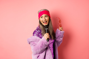 Gorgeous asian senior woman singing karaoke in mic, perform song and looking happy, standing over pink background.