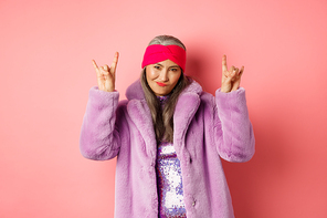 Cool asian mature woman in fashionable faux fur coat, showing rock-n-roll horns signs and smiling sassy at camera, having fun, standing over pink background.