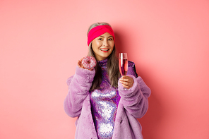 Elegant and stylish asian woman in purple faux fur coat eating donuts and drinking champagne, having fun at party, standing over pink background.