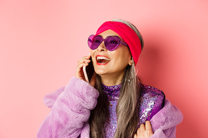 Fashion. Beautiful asian senior woman in trendy sunglasses and headband laughing, talking on mobile phone and looking aside, pink background.
