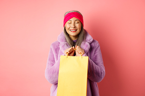Shopping and fashion concept. Happy asian senior woman in stylish clothes looking happy and delighted, standing with yellow paper bag and smiling, pink background.