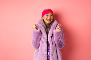 Cheerful asian lady in stylish funky coat, celebrating victory or success, saying yes and smiling happy, standing over pink background.
