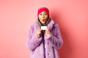 Shopping and fashion concept. Image of amazed asian senior woman checking out promo offer, holding plastic credit card and stare impressed at camera, pink background.