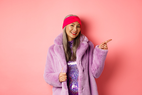 Smiling senior woman looking trendy in winter fur coat, pointing finger right and showing advertisement, standing against pink background.