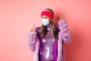 Covid-19, social distancing and fashion concept. Cheerful asian senior woman likes to party, dancing in respirator and shiny purple dress, standing over pink background.