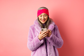 Online shopping and fashion concept. Modern asian granny in trendy faux fur coat texting message, using smartphone and looking happy at screen, pink background.