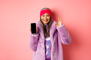 Online shopping and fashion concept. Fashionable senior asian woman showing blank black smartphone screen and OK sign, demonstrate promotion, pink background.