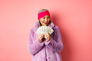 Shopping and fashion concept. Fashionable asian senior woman thinking about buying new clothes, showing money in dollars and smiling greedy, pink background.