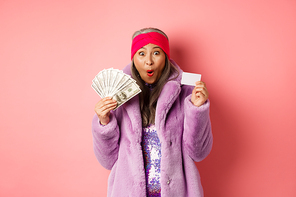 Shopping and fashion concept. Happy senior asian woman holding money in dollars and plastic card, looking amazed and ecstatic, pink background.