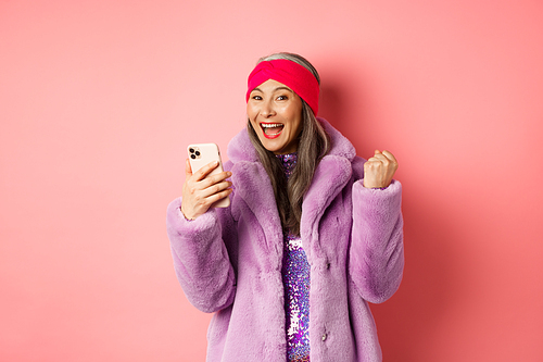 Online shopping and fashion concept. Happy asian senior woman winning prize in internet, holding mobile phone and making fist pump, scream of joy, standing over pink background.