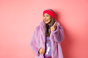 Fashion and shopping concept. Happy asian mature woman in stylish fake-fur coat pointing finger at camera, we need you gesture, standing over pin background.