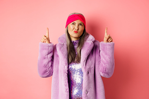 Stylish middle aged asian woman in purple fake fur coat, looking skeptical and grimacing with displeased face, pointing fingers up at something bad and unimpressive, pink background.