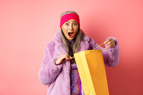 Shopping and fashion concept. Surprised asian grandmother open shop bag and looking amazed at camera, receive mother day gift, pink background.
