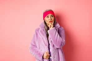 Funky old asian woman looking thoughtful and perplexed, standing in fake fur coat and thinking, making decision, standing on pink background.
