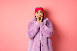Fashion concept. Trendy old asian woman looking surprised at camera. Mature female in purple coat and headband looking amazed at camera, checking out promotion on pink background.