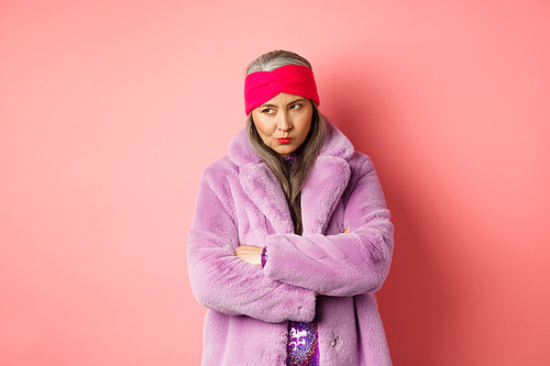 Grumpy funky old asian lady looking from under forehead, pouting and looking left with angry and disappointed face, standing in stylish purple faux fur coat against pink background.