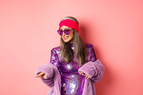 Fashionable asian senior woman dancing disco, wearing glittering shiny dress and heart-shaped sunglasses, having fun, standing over pink background.