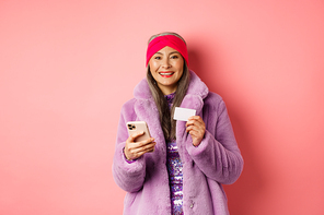 Online shopping and fashion concept. Smiling middle-aged woman in stylish fur coat using mobile phone and plastic credit card, standing happy on pink background.