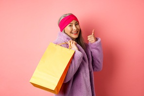 Stylish asian senior woman going shopping, carry paper bag on shoulder and showing thumb-up, recommending store discounts, pink background.
