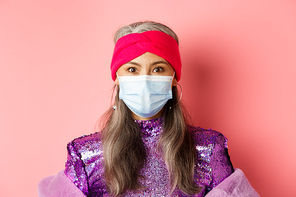 Covid-19, virus and social distancing concept. Close-up of stylish asian senior woman in medical mask and shiny dress looking at camera, standing over pink background.