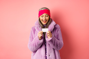Shopping and fashion concept. Beautiful and stylish asian woman looking at plastic credit card, smiling pleased, standing over pink background.