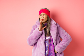 Fashion and shopping concept. Stylish asian middle-aged woman in fashionable winter clothes smell something bad, waving hand near nose from disgusting stink, pink background.