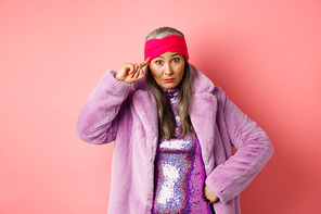Stylish asian mature woman scolding you, staring at camera annoyed and confused, pointing finger at head, are you stupid gesture, standing over pink background.