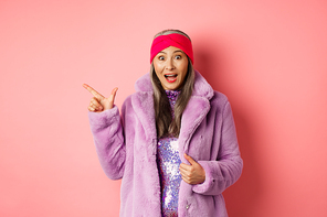 Fashion and shopping concept. Fashionable senior asian woman in winter coat and headaband pointing finger left at copy space, staring amazed at camera, checking out banner.