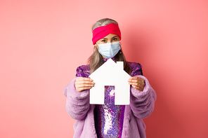 Covid-19 and real estate concept. Senior asian woman in stylish outfit and medical mask showing paper house model, searching apartment, standing over pink background.