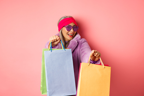 Funky and cool asian senior woman in fashionable clothes dancing while going shopping on sales, holding shop paper bags and having fun, pink background.