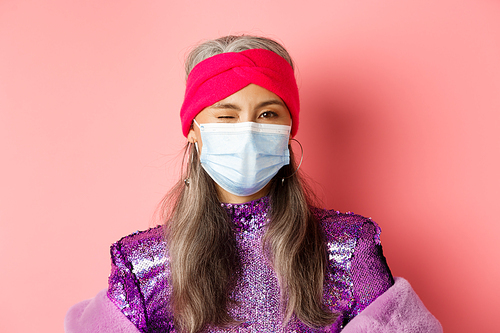 Covid-19, virus and social distancing concept. Sassy asian senior woman in medical mask and glitter dress winking, wearing party outfit, pink background.