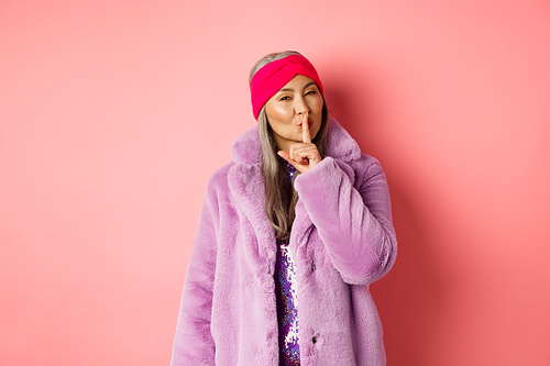 Fashion and shopping concept. Modern chinese senior woman shushing, telling a secret and looking cunning at camera, standing in trendy fake fur coat and headband on pink background.