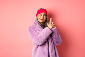 Fashion and shopping concept. Cool and stylish asian senior lady in purple fake fur coat, making finger gun gesture and looking left with sassy smile, acting like secret agent, pink background.