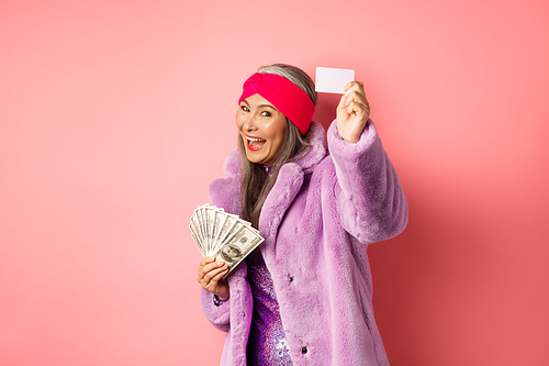 Shopping and fashion concept. Fashionable funky asian woman showing plastic credit card, paying contactless, holding money in other hand, pink background.